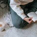 Kobieta siedzi na kocu na podłodze i czyta książkę z kawą